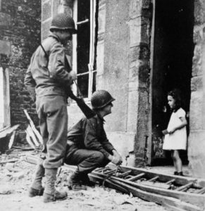 Deux soldats américains avec une petite fille sur le perron d'une maison dans le secteur de Cerisy-la-Salle le 25 juillet 1944. L'état de la cour semble indiquer que le bâtiment a souffert de bombardements. Photo : US National Archives